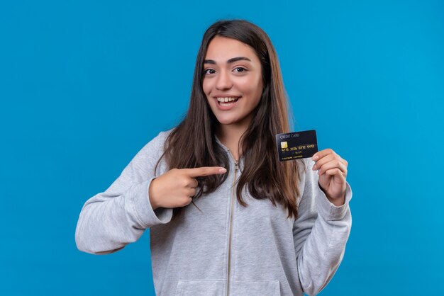 Hermosa joven con capucha gris mirando a la cámara con una sonrisa en la cara sosteniendo el teléfono y apuntando a este parado sobre fondo azul.