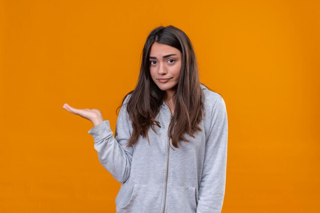 Hermosa joven con capucha gris mirando a la cámara con vista preocupada de pie sobre fondo naranja