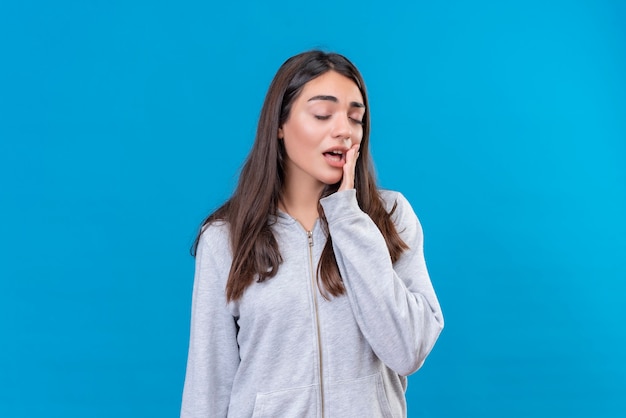 Hermosa joven con capucha gris mirando cansado sueño de pie sobre fondo azul.