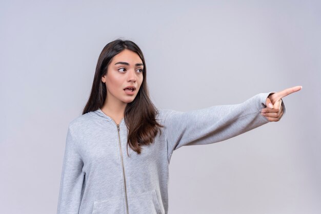 Hermosa joven con capucha gris mirando a otro lado apuntando hacia fuera de pie sobre fondo blanco.