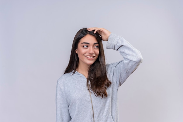 Foto gratuita hermosa joven con capucha gris mirando a otro lado y a la cámara sonríe en la cara tocando la cabeza de pie sobre fondo blanco