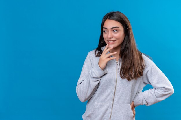 Hermosa joven con capucha gris mirando a otro lado con expresión pensativa en la cara pensando de pie sobre fondo azul.