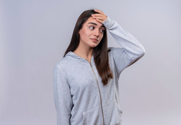 Hermosa joven con capucha gris mirando a otro lado tocando la cabeza por error mirando confundido concepto de mala memoria de pie sobre fondo blanco.