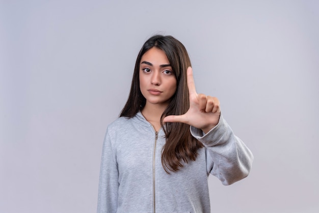 Hermosa joven con capucha gris que muestra un cartel de tamaño pequeño con un símbolo de medida de rostro serio de pie sobre fondo blanco