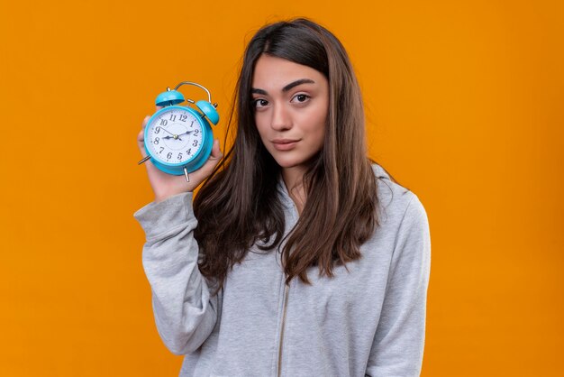 Hermosa joven con capucha gris sosteniendo el reloj y mirando a la cámara con expresión pensativa de pie sobre fondo naranja