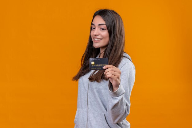 Hermosa joven con capucha gris sosteniendo tarjeta de crédito y mirando a la tarjeta de crédito con una sonrisa en la cara de pie sobre fondo naranja