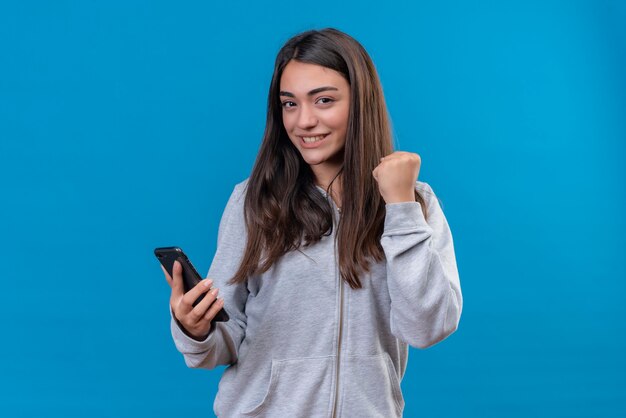 Hermosa joven con capucha gris sosteniendo el teléfono y mirando a la cámara con la cara existente de pie sobre fondo azul.