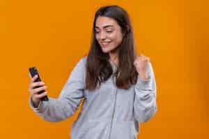 Foto gratuita hermosa joven con capucha gris sosteniendo el teléfono con una sonrisa en la cara de pie sobre fondo naranja