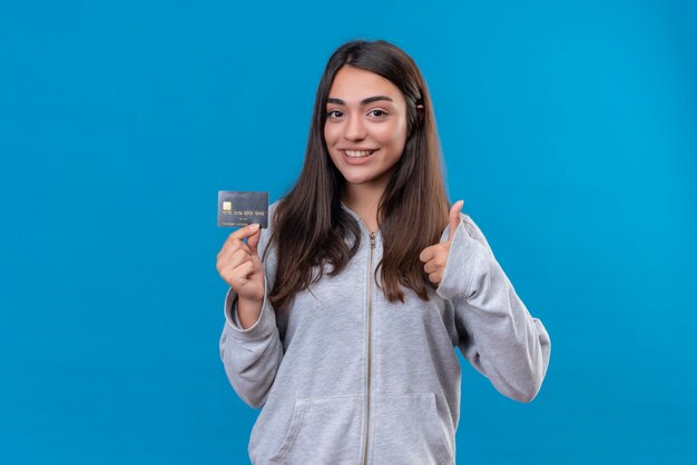 Hermosa joven con capucha gris con tarjeta de crédito y mirando a cámara con una sonrisa en la cara y hacer como gesto de pie sobre fondo azul.