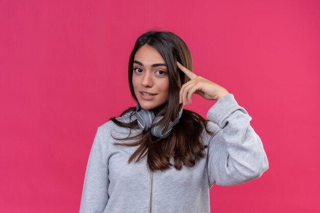 Hermosa joven con capucha gris usando audífonos mirando a la cámara tocando la cabeza con el dedo de pie sobre fondo rosa