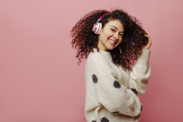 Hermosa joven caucásica con cabello rizado escuchando música con auriculares sobre fondo rosa en el estudio Morena fuma y usa suéter blanco cálido Concepto de belleza y juventud