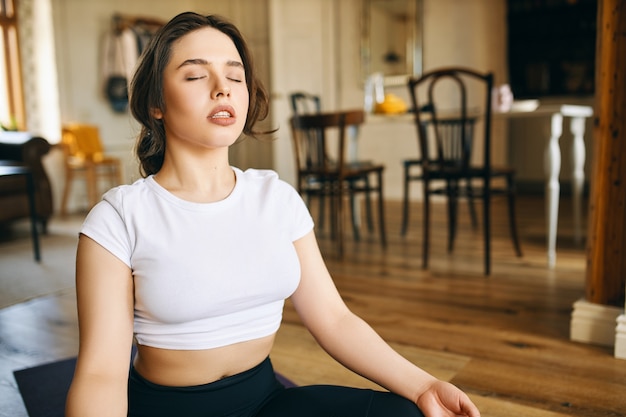 Hermosa joven caucásica con cuerpo musculoso y curvilíneo sentado en postura de loto en casa manteniendo los ojos cerrados, meditando durante la práctica de yoga, haciendo escaneo corporal, concentrándose en la respiración