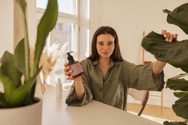 Hermosa joven caucásica con tarro de mascarilla para el cabello sentada en la mesa en el interior Morena con camisa verde mira a la cámara Concepto de belleza y cuidado