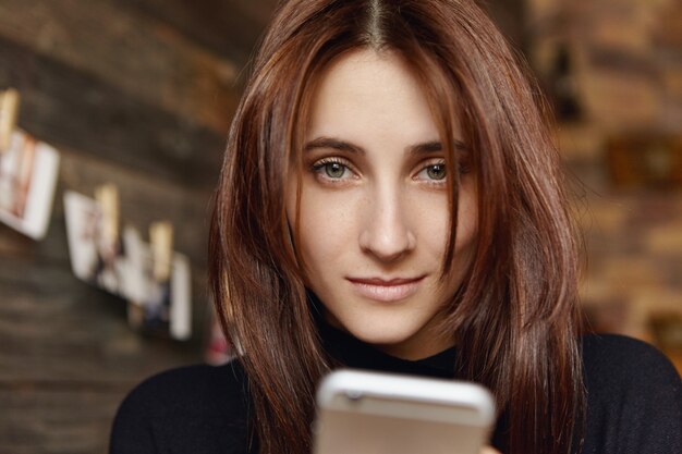 Hermosa joven caucásica wirh cabello oscuro con teléfono celular, mensajes a amigos en línea o navegar por internet sentado en el interior de la cafetería moderna. Concepto de personas, tecnología y comunicación.