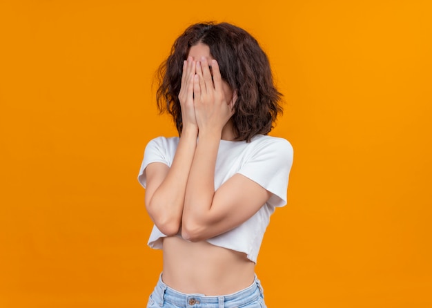 Hermosa joven cerrando su rostro con las manos sobre fondo naranja aislado con espacio de copia