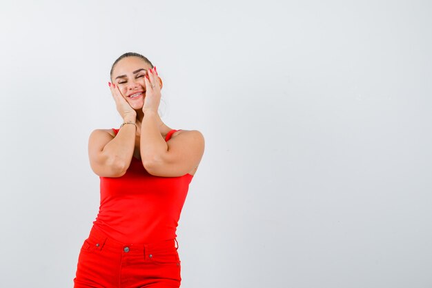 Hermosa joven cogidos de la mano en las mejillas en camiseta roja, pantalones y mirando feliz. vista frontal.