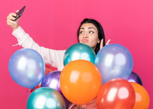 La hermosa joven complacida de pie detrás de los globos toma un selfie mostrando un gesto de paz aislado en la pared rosa