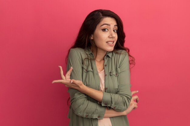 Hermosa joven confundida con camiseta verde oliva aislada en la pared rosa con espacio de copia