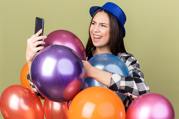 Hermosa joven confundida con sombrero azul de pie detrás de globos sosteniendo y mirando el teléfono aislado en la pared verde oliva