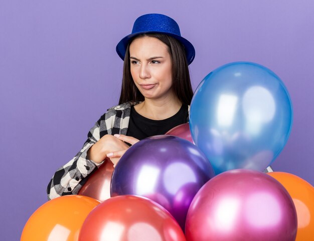 Hermosa joven confundida con sombrero de fiesta de pie detrás de globos