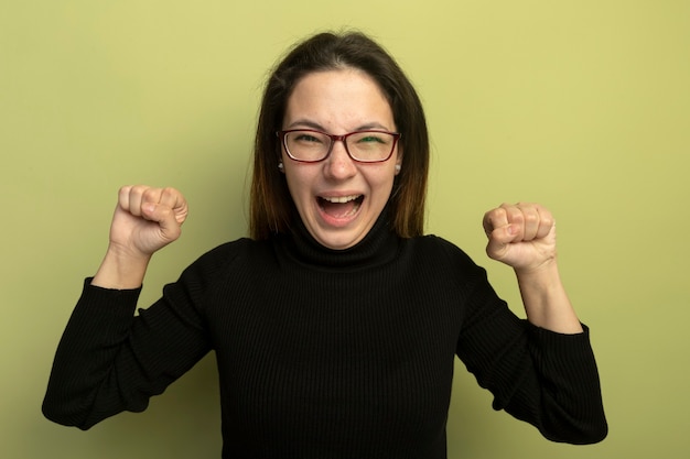 Hermosa joven en un cuello alto negro y gafas apretando los puños feliz y emocionado