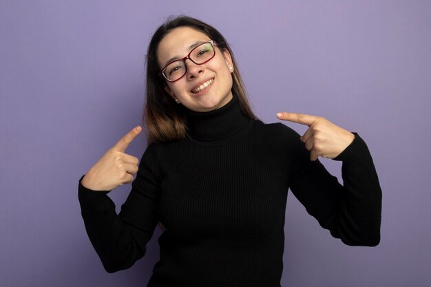 Hermosa joven en un cuello alto negro y gafas apuntando con los dedos a sí misma feliz y positiva