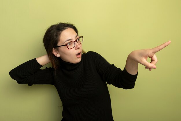 Hermosa joven en un cuello alto negro y gafas apuntando con el índice figner a algo que se sorprende