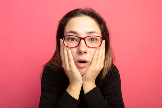 Hermosa joven en un cuello alto negro y gafas asombrado y sorprendido