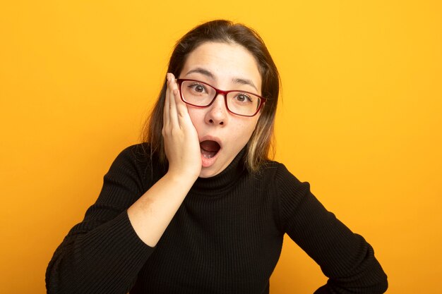 Hermosa joven en un cuello alto negro y gafas asombrado y sorprendido