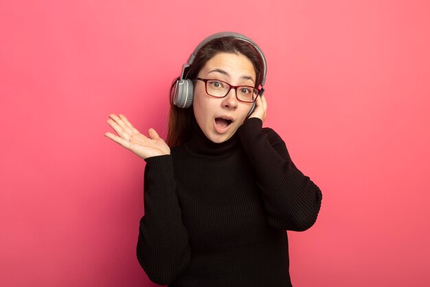 Hermosa joven en un cuello alto negro y gafas con auriculares feliz y positivo con el brazo levantado