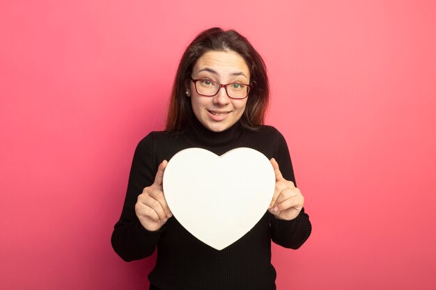 Hermosa joven en un cuello alto negro y gafas con caja en forma de corazón sonriendo