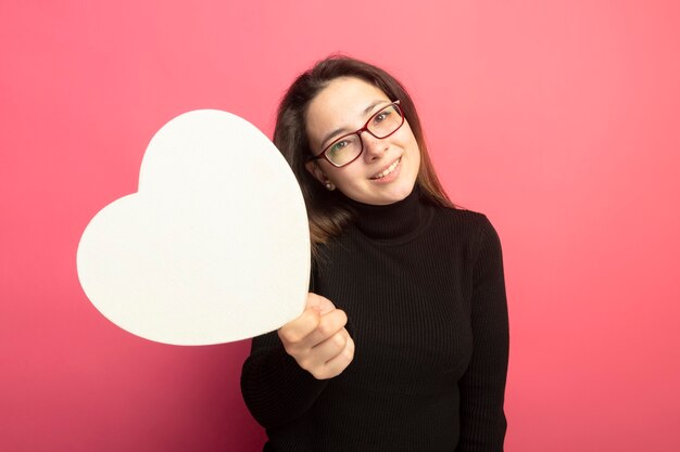 Hermosa joven en un cuello alto negro y gafas con corazón de cartón sonriendo con cara feliz