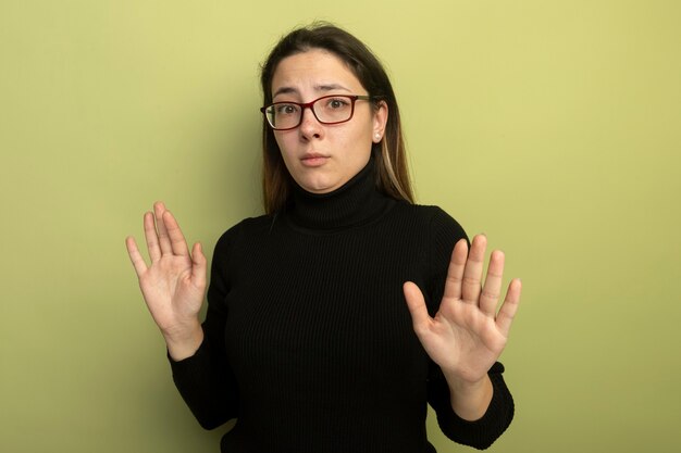 Hermosa joven en un cuello alto negro y gafas con expresión de miedo sosteniendo las manos