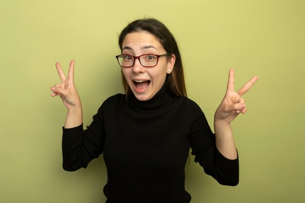 Hermosa joven en un cuello alto negro y gafas, feliz y emocionada mostrando v-sign