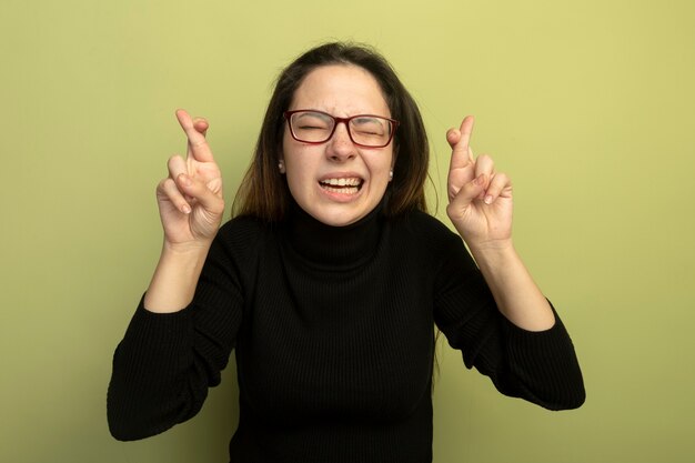 Hermosa joven en un cuello alto negro y gafas haciendo deseo deseable cruzando los dedos con expresión de esperanza con los ojos cerrados