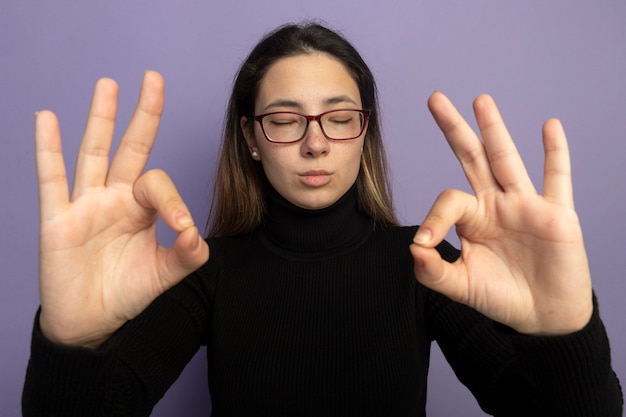 Hermosa joven en un cuello alto negro y gafas haciendo gesto de meditación con los dedos con los ojos cerrados