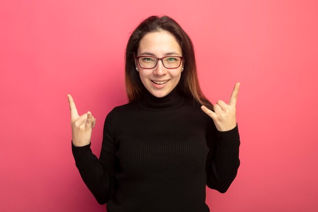 Hermosa joven en un cuello alto negro y gafas lookign a la cámara sonriendo alegremente haciendo símbolo de rock