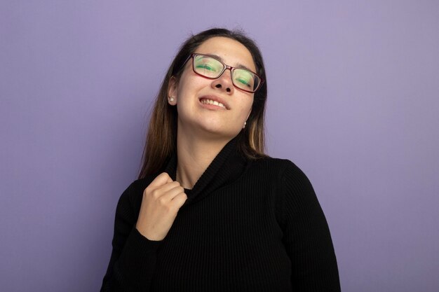 Hermosa joven con un cuello alto negro y gafas mirando a la cámara con expresión molesta tocando su cuello