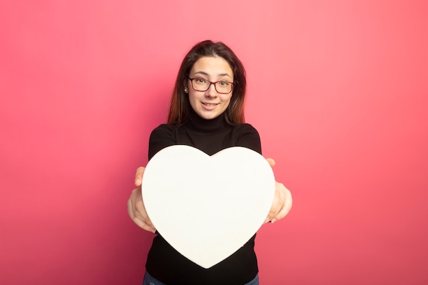 Hermosa joven en un cuello alto negro y gafas mostrando caja en forma de corazón con una sonrisa en la cara