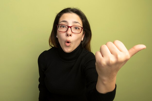 Hermosa joven en un cuello alto negro y gafas sorprendieron apuntando con el pulgar hacia un lado