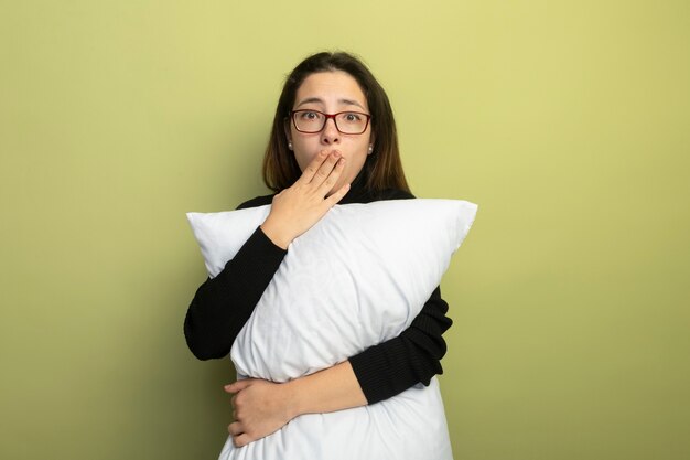 Hermosa joven en un cuello alto negro y gafas sosteniendo la almohada conmocionado cubriendo la boca con la mano