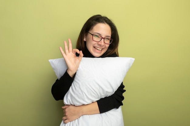 Hermosa joven en un cuello alto negro y gafas sosteniendo la almohada sonriendo alegremente mostrando el signo de ok