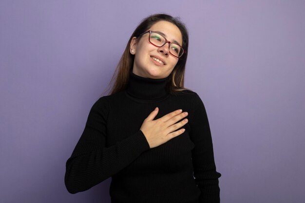 Hermosa joven en un cuello alto negro y gafas sosteniendo la mano sobre su pecho sonriendo sintiéndose agradecido