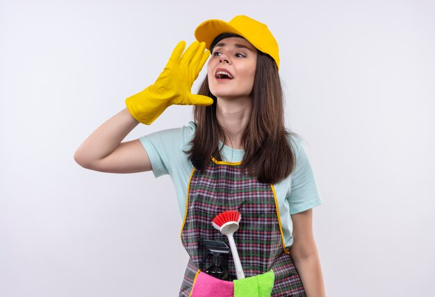 Hermosa joven con delantal, gorra y guantes de goma gritando o llamando con la mano cerca de la boca