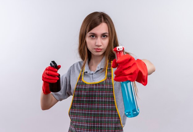 Hermosa joven en delantal y guantes de goma con aerosoles de limpieza apuntando con ellos con cara seria