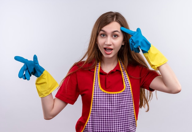 Hermosa joven en delantal y guantes de goma apuntando con el dedo índice hacia el lado mirando sorprendido