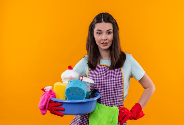 Hermosa joven en delantal y guantes de goma con lavabo con herramientas de limpieza mirando a la cámara con expresión segura