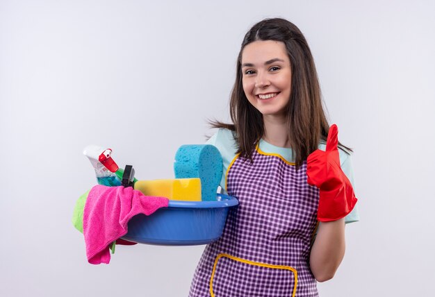 Hermosa joven en delantal y guantes de goma con lavabo con herramientas de limpieza sonriendo con cara feliz mostrando los pulgares para arriba