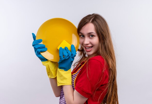 Hermosa joven en delantal y guantes de goma lavando placa con esponja, sonriendo alegremente