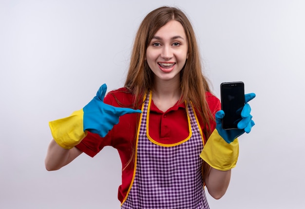 Hermosa joven en delantal y guantes de goma mostrando smartphone apuntando con el dedo sonriendo alegremente